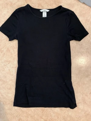 Svart ribbad t-shirt från H&M - En enkel svart ribbad t-shirt från H&M med rund halsringning och korta ärmar. T-shirten har en smal passform och är tillverkad i ett mjukt, stretchigt material som sitter skönt på kroppen. Perfekt basplagg till garderoben.