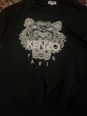 Svart sweatshirt med tiger från Kenzo - Svart sweatshirt från Kenzo med ikonisk broderad tiger och texten 'KENZO PARIS' i vitt framtill. Tröjan har rund halsringning och ribbade muddar. Perfekt för dig som gillar streetwear och vill sticka ut med en klassisk design.