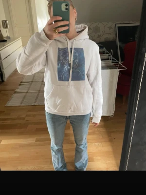 Vit hoodie med konstnärligt tryck - Säljer en vit hoodie med stor huva och dragsnören. På bröstet finns ett blått konstnärligt tryck av ett ansikte i linjekonst. Hoodien har en klassisk känguruficka framtill och är gjord i mjukt material, perfekt för en chill och avslappnad stil.