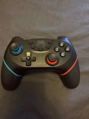 Svart trådlös spelkontroll Switch - Snygg svart trådlös handkontroll till Nintendo Switch med färgdetaljer i blått och rött runt styrspakarna. Ergonomisk design med gummerade handtag för bättre grepp och klassiska knappar som X, Y, A, B samt styrkors.