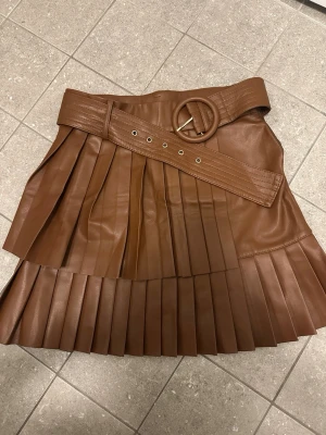 Brun plisserad kjol från Zara - Säljer en brun plisserad kjol från Zara i skinnimitation. Kjolen har ett brett bälte med rund spänne i midjan och snygga detaljer som ger en cool och trendig look. Perfekt för dig som vill sticka ut lite extra.