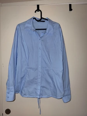 Ljusblå skjorta med snörning bak - Stilren ljusblå skjorta i bomull med klassisk krage och knappar framtill. Unik detalj med snörning i ryggen för att få en mer figursydd look! (Går att dra ihop olika hårt beroende på hur tight man vill ha den)