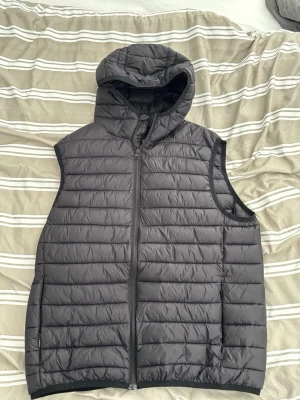 Svart dunväst med huva - Snygg svart dunväst med huva och dragkedja framtill. Västen har quiltad design med horisontella sömmar och är lätt vadderad för extra värme. Perfekt att slänga över en hoodie eller tröja för en chill och stilren look.