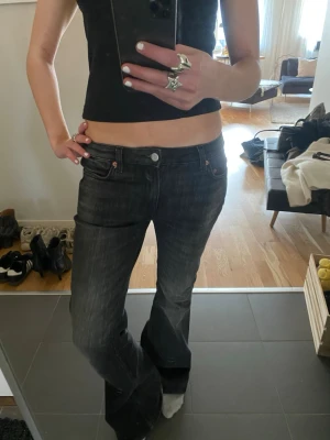 Svarta bootcut jeans med låg midja - Säljer ett par grå/svarta bootcut jeans med låg midja och klassisk femficksmodell. Jeansen har en lätt tvättad look och vida ben som ger en snygg retrostil. Perfekta för dig som gillar en avslappnad men trendig vibe.