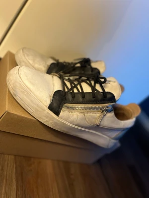 Giuseppe zanotti skor - Säljer ett par vita sneakers från Giuseppe Zanotti med svarta detaljer, dragkedja på sidan och svarta skosnören. Skorna har en låg profil och är tillverkade i läder. Blixtsymbol på plösen ger en unik touch. Perfekta för dig som gillar streetstyle.