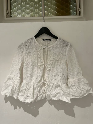 Vit broderad blus från Zara - Söt vit blus från Zara med broderie anglaise-mönster och volang nedtill. Blusen har långa ärmar med lätt puff och knytband framtill. Tillverkad i bomull för en luftig och romantisk känsla.