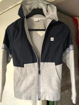 Grå och marinblå hoodie från BOSS - Snygg hoodie från BOSS i grått och marinblått med dragkedja framtill och huva. Har BOSS-logga på bröstet och band med BOSS-tryck längs ärmarna. Tillverkad i mjukt sweatshirtmaterial och med fickor fram. Perfekt för en avslappnad och trendig stil. Passar till 12 år