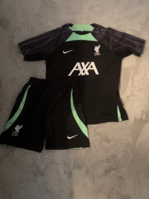 Liverpool träningsset Nike svart/grön - Säljer ett Liverpool FC träningsset från Nike. Setet består av en svart t-shirt och shorts med gröna detaljer och vita loggor. AXA och LFC-logga på bröstet samt Nike swoosh. Perfekt för fotbollsträning eller gymmet.