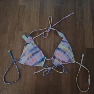 Färgglad bikiniöverdel med knyt - Säljer en snygg trekantsbikini med knyt i nacken och ryggen. Bikinin har ett färgglatt mönster i pastelliga toner av rosa, blått, gult och lila. Perfekt för stranden eller poolen och ger en riktigt somrig vibe.