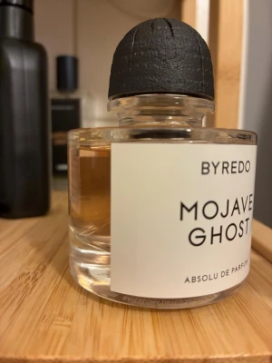 Byredo Mojave Ghost Absolu de Parfum - Endast testad några gånger. Byredo Mojave Ghost Absolu de Parfum i en stilren, rund glasflaska med svart kork. Doften har en ljus bärnstensfärgad vätska och etiketten är vit med svart text. Flaskan känns lyxig och modern, perfekt för dig som gillar exklusiva parfymer.