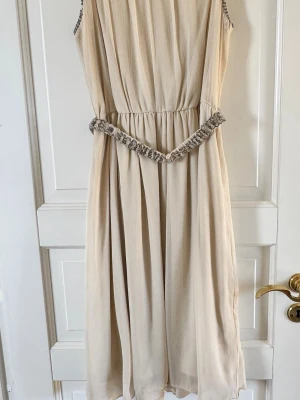 Beige plisserad klänning med pärldetaljer - Elegant beige klänning från KappAhl i plisserat tyg med pärlbroderade detaljer vid halsringning och midja. Klänningen är ärmlös och har ett mjukt fall som ger en romantisk look. Perfekt för dig som vill ha en stilren och feminin outfit.