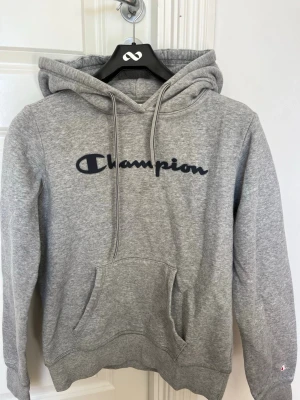 Grå hoodie från Champion - Snygg grå hoodie från Champion i storlek M med stor logga i svart på bröstet. Använd ett få antal gånger, likadant med tvätt. Den sitter normalt. Passar perfekt till att gå ut, men också bara vara avslappnad hemma.