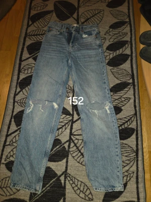 Blå raka jeans med slitningar - Säljer ett par blå raka jeans med slitningar över knäna. Jeansen har klassisk femficksdesign och hög midja. Perfekta för en avslappnad och trendig look.