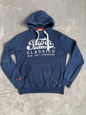 Marinblå Superdry Hoodie - Säljer en riktigt fet hoodie från superdry! Hoodien är en indigo edition, vilket betyder att den har en speciell indigo blå färg och ska passa bättre till jeans. Är i mycket fint skick! Storlek M Hör av dig vid minsta fundering!😊