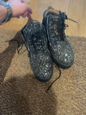 Svarta glittriga giuseppe zanotti med dragkedja - Säljer ett par giuseppe zanotti sneakers täckta med glitter och med svarta snören. Skorna har dragkedjor på sidorna och svart sula. Insidan är beige och materialet ser ut att vara textil med glitterdetaljer. Perfekta för dig som vill sticka ut! För mer bilder/videos kom dm