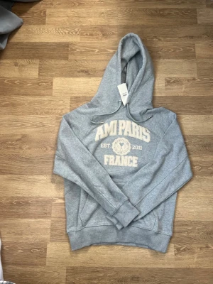 Grå hoodie från AMI Paris - Snygg grå hoodie från AMI Paris med stor vit text framtill och tryck med 'AMI PARIS FRANCE EST 2011'. Klassisk huva med dragsko och ribbade muddar. Tillverkad i mjuk bomullsblandning, perfekt för en avslappnad streetwear-look.