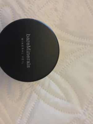 bareMinerals Mineral Veil Foundation - Säljer en bareMinerals Mineral Veil foundation i burk. Produkten är i lös puderform och har en lätt tonad nyans. Förpackningen är rund och svart med transparent botten som visar pudrets färg. Perfekt för en naturlig finish och matt look.
