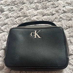 Svart handväska från Calvin Klein - Snygg svart handväska från Calvin Klein i slätt konstläder. Väskan har en stilren, rektangulär form med silverfärgad CK-logga framtill och matchande dragkedjor. Praktiskt handtag upptill gör den enkel att bära. Perfekt för dig som gillar minimalistisk och modern design.