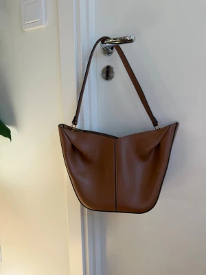 Snygg och populär brun axelremsväska  - Snygg handväska i brun läderimitation med enkel axelrem och mjuk, rundad form. Väskan har minimalistisk design med synliga sömmar framtill och guldfärgade detaljer vid remfästena. Perfekt för dig som gillar stilrena accessoarer.