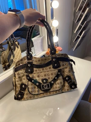 Beige Guess handväska med detaljer - Säljer en beige handväska från Guess med bruna och svarta detaljer, silverfärgade nitar och spännen. Väskan har ett mönster med G-loggor över hela ytan och ett dekorativt spänne framtill. Handtagen är i mörkt skinnliknande material.