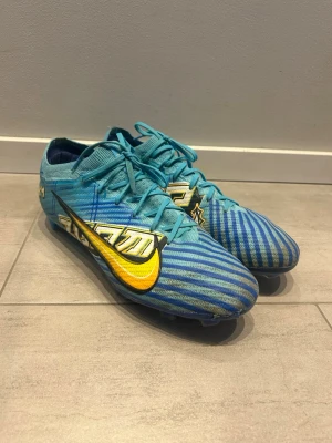 Nike Mercurial Air Zoom - Säljer ett par Nike Mercurial fotbollsskor i blått med gula och vita detaljer samt ett stort gult Nike-märke på sidan. Skorna har snörning och är tillverkade i ett lätt syntetmaterial med mönstrad ovandel för bättre bollkontroll. Perfekta för dig som vill sticka ut på planen. Dom är lite slitna på utsidan och det finns inga sulor till skorna!!