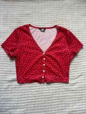 Röd croppad topp med hjärtan - Söt croppad topp i röd färg med små vita hjärtan, v-ringad och med knappar framtill. Kortärmad och i mjukt ribbat material, perfekt för en trendig och avslappnad look. Aldrig använd!💓🌸
