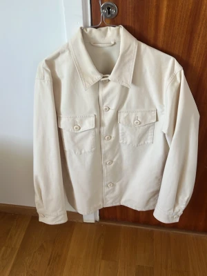 Beige overshirt med fickor fram - Stilren beige overshirt i bomull med klassisk krage och knäppning framtill. Två bröstfickor med lock och knapp för en clean look. Perfekt att bära öppen eller stängd över t-shirt. Långärmad och rak passform.