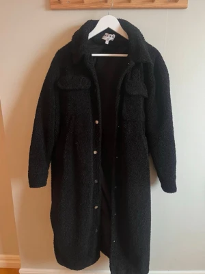 Svart lång teddyjacka från NA-KD - Säljer en svart lång teddyjacka från NA-KD med stora bröstfickor och krage. Jackan har knappar framtill och är gjord i mjukt, fluffigt teddy-material som håller dig varm och stylish. Perfekt för kalla dagar och enkel att matcha med olika outfits.