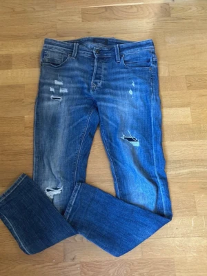 Jack & Jones jeans med slitningar  - Säljer ett par blå jeans med slitningar och lagade detaljer på både lår och knä. Jeansen har klassisk femficksmodell, normal midja och raka ben. Materialet är denim i bomull och de har en cool, avslappnad look.