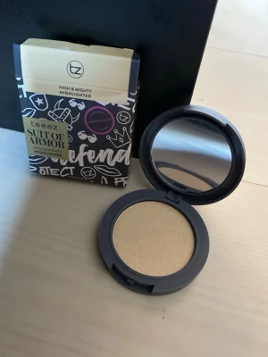 Teeez Suit of Armor Highlighter - Highlighter från Teeez i nyansen Suit of Armor. Kommer i en rund dosa med spegel och har en skimrande guldig ton. Förpackningen är svart med grafiska vita och guldiga detaljer samt text. Perfekt för att ge glow till ansiktet.