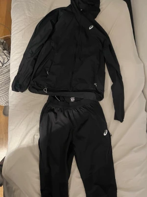 Svart träningsset från Asics - Säljer ett svart träningsset från Asics med jacka och byxor. Jackan har huva, dragkedja och logga på bröstet. Byxorna har resår i midjan och Asics-logga på benet. Perfekt för löpning eller annan träning utomhus.