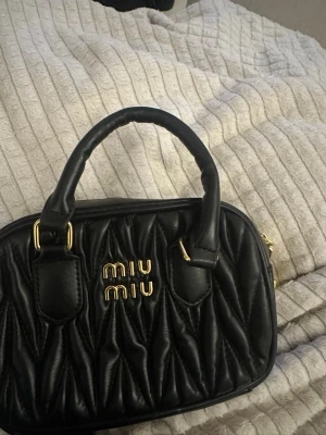 Svart quiltad handväska från Miu Miu - Snygg svart quiltad handväska från Miu Miu med guldfärgade detaljer och logga framtill. Väskan har två handtag och avtagbar axelrem i svart läder. Perfekt storlek för det viktigaste och ger en lyxig känsla till din outfit. Köptes i tokyo secondhand och säljer vidare för mycket billigare! pris går definitivt att diskutera ❣️