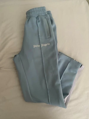 Ljusblå byxdress från Palm Angels - Snygg ljusblå byxdress från Palm Angels med vita och svarta ränder längs sidorna. Byxdressen har resår i midjan och raka ben. Tillverkad i ett mjukt och följsamt material som ger en sportig vibe. Perfekt för dig som gillar streetwear och vill sticka ut.  OBS! Har många fläckar! 