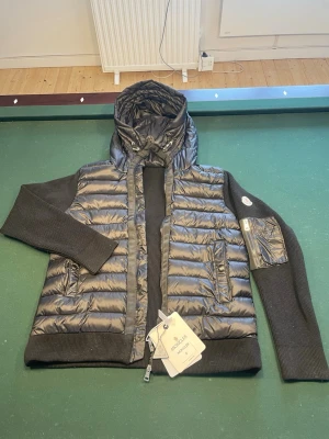 Moncler cardigan - Snygg svart cardigan från Moncler med stickade svarta ärmar och huva. Den har två framfickor med knappar, dragkedja framtill och en Moncler-logga på ärmen. Materialet är en kombination av dun och stickad textil för en modern look. Den är i väldigt bra skick och priset kan diskuteras 
