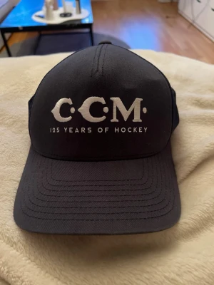 Svart keps från CCM 125 Years of Hockey - Snygg svart keps från CCM med vit brodyr framtill där det står '125 Years of Hockey'. Klassisk trucker-modell med böjd skärm och mesh på sidorna för extra ventilation. Justerbar baktill för perfekt passform.