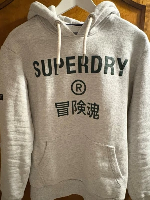 Grå hoodie från Superdry - Snygg grå hoodie från Superdry med svart logga och japanska tecken på bröstet. Hoodien har huva med vita dragsnören och en stor magficka. Tillverkad i mjukt och bekvämt material, perfekt för chill dagar eller när du vill ha en avslappnad stil. Storlek S men passar även M suveränt