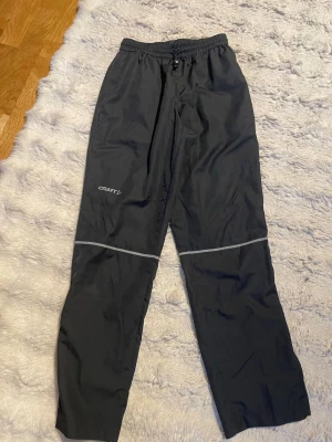 Svarta täckbyxor från Craft - Svarta täckbyxor från Craft med elastisk midja och dragsko. Byxorna har reflexdetaljer runt knäna och är tillverkade i ett lätt, vind- och vattenavvisande material. Perfekta för kalla dagar och utomhusaktiviteter. Snygg och sportig design.