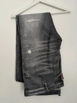 Grå slitna jeans från Dsquared2 - Säljer ett par grå jeans från Dsquared2 med coola slitningar, färgstänk och distressed detaljer. Jeansen har klassisk femficksmodell och synlig röd logotagg vid fickan. Perfekta för en avslappnad streetwear-look.