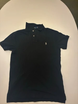 Svart pikétröja från Ralph Lauren - Klassisk svart pikétröja från Ralph Lauren med vit broderad logga på bröstet. Tröjan har korta ärmar, krage och två knappar framtill. Perfekt för en clean och stilren look.