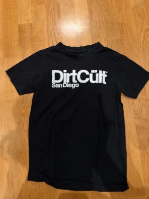 Svart DirtCült t-shirt San Diego - Säljer en svart t-shirt från DirtCült med stor vit logga och texten 'San Diego' på bröstet. Klassisk rak passform och rund halsringning. Perfekt för dig som gillar streetwear och vill ha en enkel men snygg look.