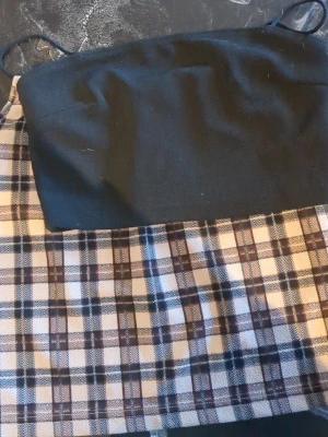 Rutig bandeau topp med svarta detaljer - Trendig bandeau topp med rutigt mönster i beige, svart och brunt. Överdelen är helsvart och har tunna axelband. Perfekt för dig som vill ha en cool och enkel look till sommaren.