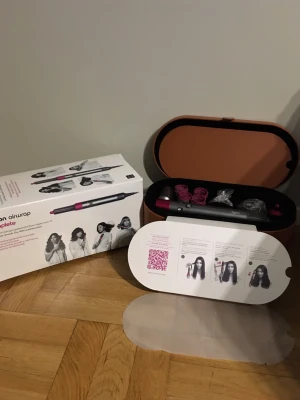 Dyson Airwrap Complete hårstyler - Säljer en Dyson Airwrap Complete hårstyler i grå och rosa med flera tillbehör för styling. Kommer i en snygg brun förvaringsväska och originalkartong. Perfekt för att skapa lockar, volym och släta frisyrer. Inkluderar olika borsthuvuden och instruktioner.