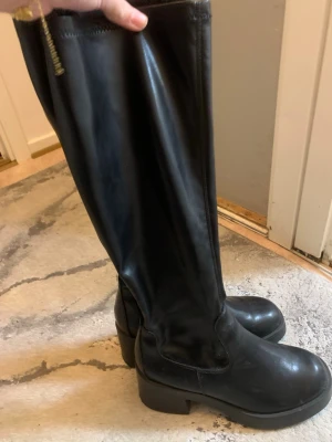 Svarta knähöga läderstövlar - Säljer ett par svarta knähöga stövlar i slätt läder med dragkedja på insidan. De har en chunky klack och rundad tå, vilket ger en cool och tidlös look. Perfekta till jeans eller kjol för en trendig outfit.