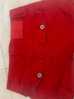 Röda jeans med stora fickor - Snygga röda jeans med stora fickor framtill och bak, samt dekorativa knappar. Jeansen har breda hällor och dragkedja. Materialet är kraftig denim i bomull. Perfekta för dig som vill sticka ut med färg och cool design.