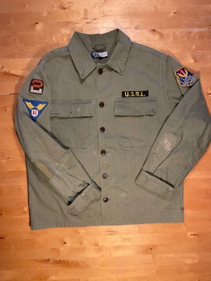 Grön Ralph Lauren overshirt med patchar - Säljer en militärinspirerad grön overshirt från Rothco med coola patchar på ärmar och bröst, bland annat U.S.R.L. och färgglada märken. Skjortan har två bröstfickor med lock, knäppning framtill och klassisk krage. Tillverkad i slitstark bomull.