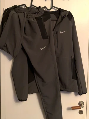 Grå Nike träningsset med huva - Säljer ett grått träningsset från Nike med matchande byxor och jacka. Jackan har huva, dragkedja och svarta detaljer samt Nike-logga på bröstet. Byxorna har resår i midjan och smal passform. Perfekt för träning eller chill.
