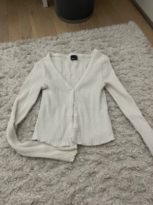Vit ribbad kofta från Gina Tricot - Säljer en vit ribbad kofta från Gina Tricot med v-ringning och knappar framtill. Koftan har långa ärmar och är tillverkad i ett mjukt, stretchigt material som sitter snyggt på kroppen. Perfekt att styla med jeans eller kjol.