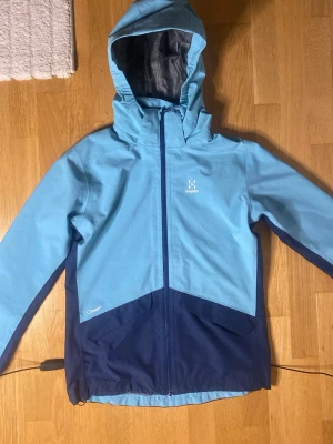 Blå regnjacka från Haglöfs - Snygg tvåfärgad regnjacka från Haglöfs i ljusblått och marinblått. Jackan har huva, hel dragkedja framtill och är tillverkad i ett vattenavvisande material. Perfekt för regniga dagar och outdoor-äventyr. Diskret Haglöfs-logga på bröstet och elastiska muddar.