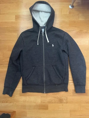 Ralph lauren zip-hoodia - Säljer nu en riktigt fet hoodie från ralph lauren. Den har vita drawstrings och snygg svart/mörkgrå färg. Den är i lite tjockare mer premiummaterial än andra ralph lauren hoodies och är därför perfekt nu på våren eller för kyliga sommarkvällar. Storlek S, passar även M. Inga större defekter eller skavanker utan den är i väldigt gott skick. Tveka ej att skriva vid funderingar eller liknande!