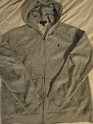 Grå hoodie från Polo Ralph Lauren - Snygg grå hoodie från Polo Ralph Lauren med dragkedja framtill och klassisk huva. Tröjan har två fickor och den ikoniska lilla loggan broderad på bröstet. Tillverkad i mjukt bomullsmaterial som är skönt att ha på sig.